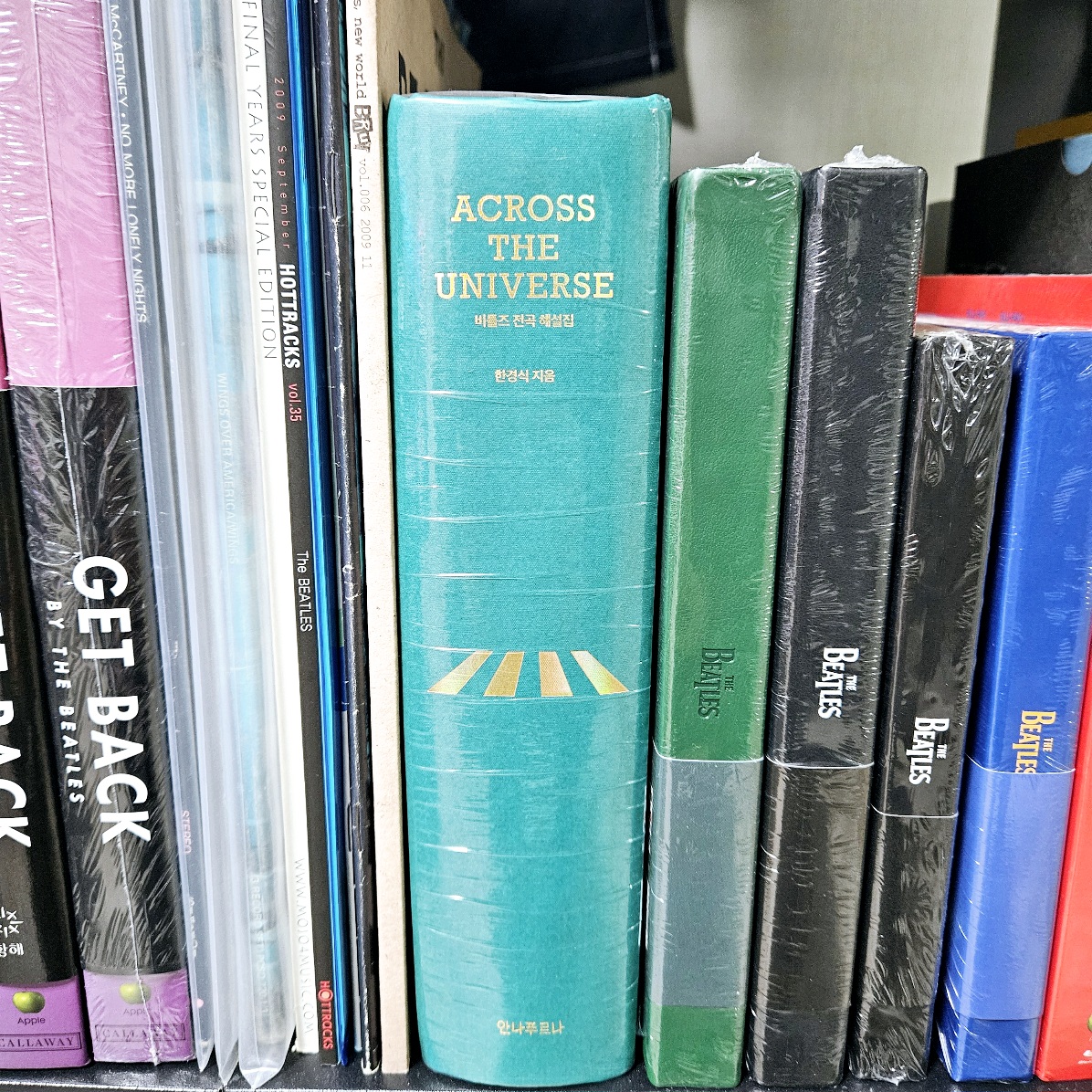 초록색 하드커버로 제작된 '우주를 가로지르는 비틀즈 전곡 해설집'이 책장에 꽂혀 있는 모습. 금박으로 인쇄된 'Across The Universe' 타이틀이 돋보이며, 주변에는 비틀즈 관련 CD와 책들이 함께 진열되어 있다.
