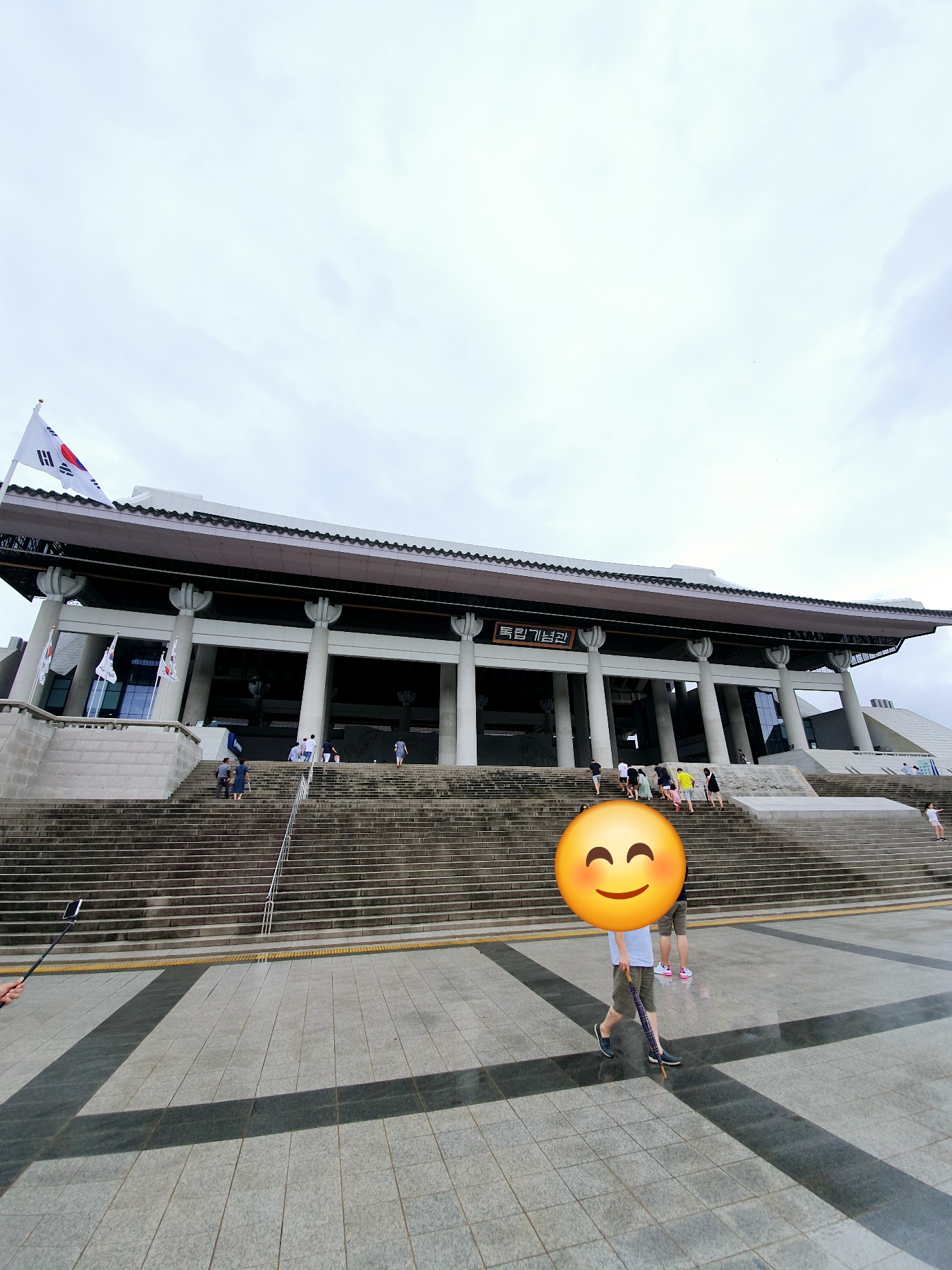 독립기념관 본관 외부 계단과 전경을 배경으로 방문객이 걸어가는 장면. 하늘이 열린 광장과 태극기가 함께 보이는 넓은 공간 구성.