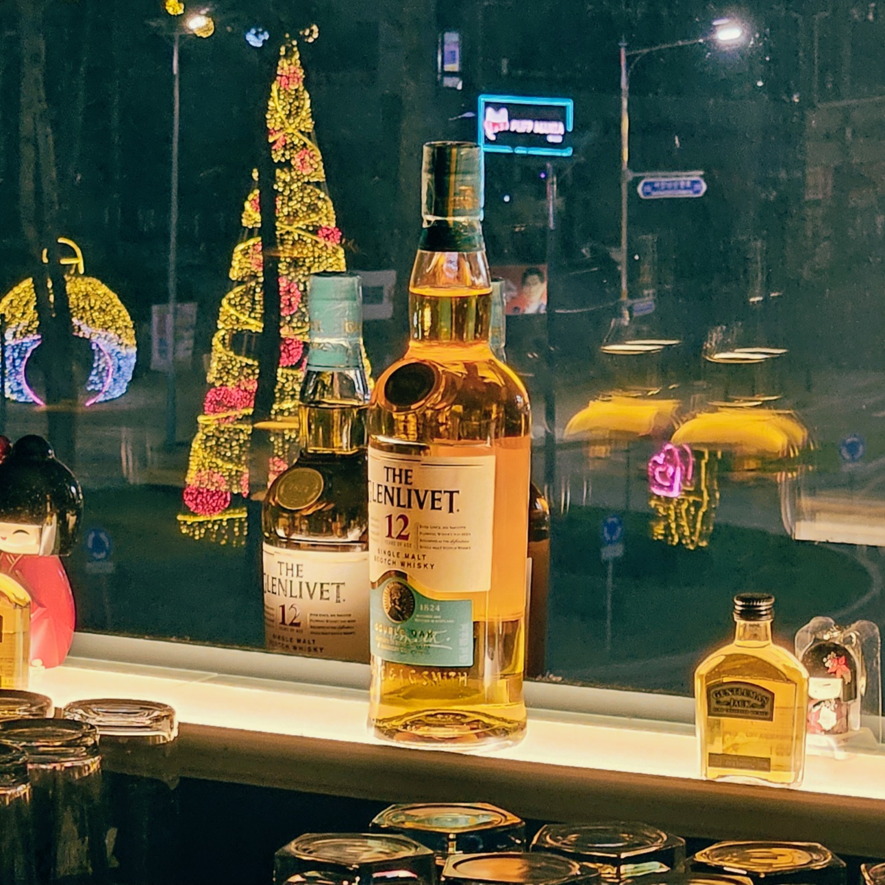야간 창가에 전시된 글렌리벳 12년 위스키 병. 유리창에 비친 조명과 어우러져 황금빛 위스키가 은은하게 반짝인다.